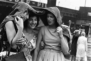 Das Foto von Pressefotografin Marga Kingler zeigt drei fröhliche Frauen vor dem Essener Hauptbahnhof, die gemeinsam Schutz unter einer Jacke vor dem Sommerregen im August 1961 in Essen suchen.