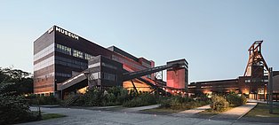 Außenansicht des Ruhr Museums auf dem UNESCO Welterbe Zollverein Moderne Außenansicht des Ruhr Museums auf dem Gelände des UNESCO-Welterbes Zeche Zollverein in Essen bei Tageslicht. Das Gebäude besteht aus dunklen Ziegel- und Stahlstrukturen mit industriellem Charakter, betont durch gläserne Fensterbänder und markante Treppenanlagen. Ein beleuchteter Förderturm sowie beleuchtete Laufgänge und Wege ergänzen das Ensemble.
