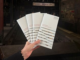 Das neue Winter-Halbjahresprogramm des Ruhr Museums Eine Hand hält vier aufgefächerte Exemplare des neuen Veranstaltungsprogramms des Ruhr Museums für Oktober 2026 bis März 2027. Im Hintergrund ist unscharf der Eingangsbereich des Ruhr Museums zu sehen.
